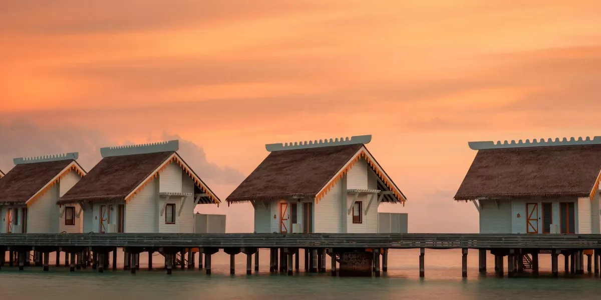 Thatched-roof overwater villas on stilts against an orange sunset sky