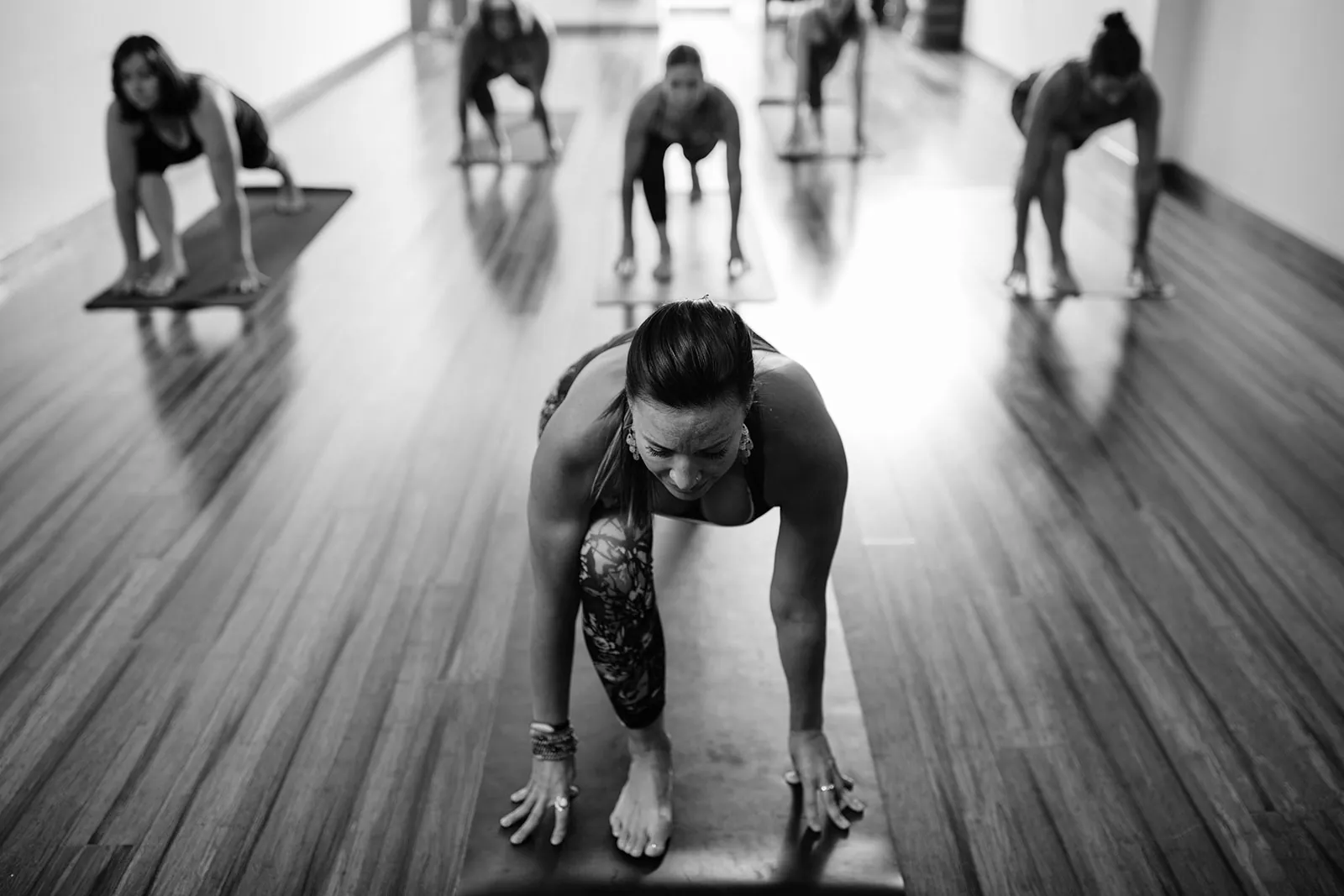 Black and white view of an instructor in a lunge with hands on the mat and students behind on a wood floor