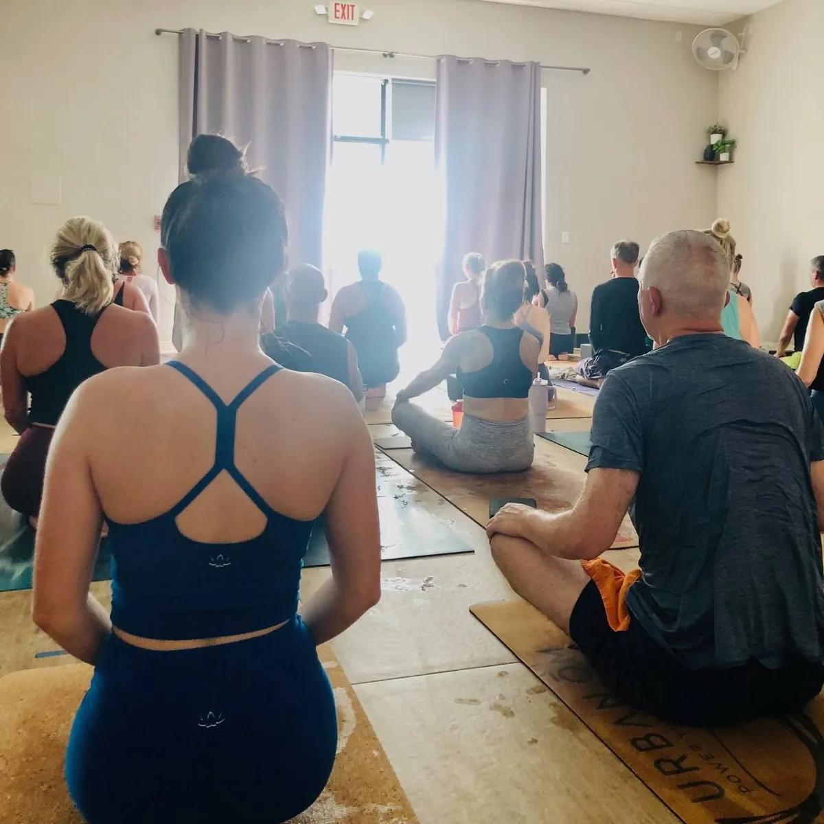 Class seated on mats facing a sunlit window in the studio, with an Urban Ōm cork mat in the corner