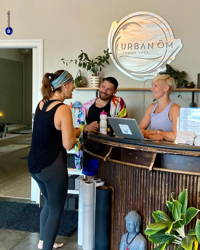 Three people chatting at the Urban Ōm Power Yoga front desk in Heathrow beside a Buddha statue and plants