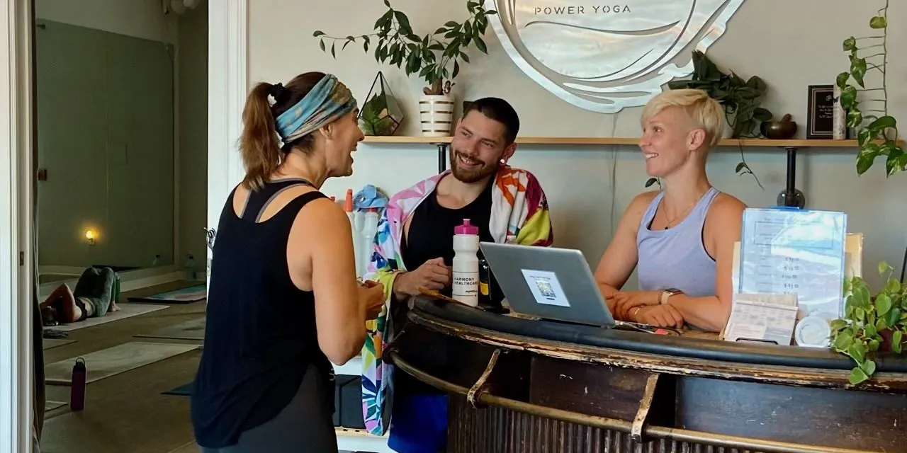 Three people chatting and smiling at the Urban Ōm Power Yoga front desk in Heathrow, under the studio sign