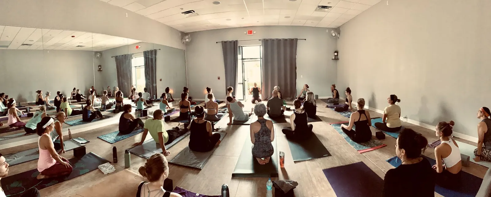 Wide view of a full yoga studio with a mirrored wall and students seated on mats around a person at the front